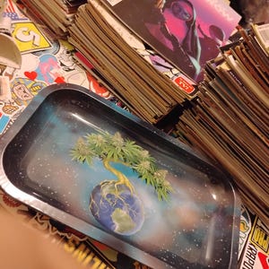 May include: A rectangular metal rolling tray with a blue and white space-themed background. The tray features a tree growing out of a globe. A stack of comic books is visible in the background.