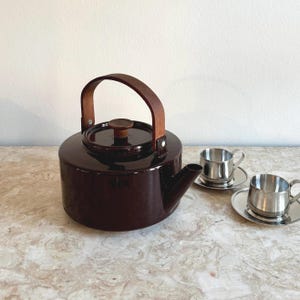 Peut inclure: Une théière en céramique marron foncé avec une poignée et un couvercle en bois, accompagnée de deux petites tasses et soucoupes en acier inoxydable. La théière a une forme arrondie et un bec verseur. Les tasses sont petites et brillantes.