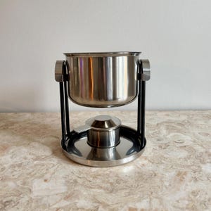 Può includere: Una pentola per fonduta in acciaio inossidabile con un supporto in metallo nero. La pentola ha una forma a ciotola arrotondata e si trova sopra un piccolo bruciatore e una base circolare. Il supporto ha una maniglia su ogni lato della pentola.