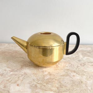 Könnte beinhalten: Eine goldfarbene Teekanne mit schwarzem Griff. Die Teekanne hat einen abgerundeten Körper, einen langen Ausguss und einen Deckel mit einer kleinen Öffnung. Der Griff ist ein gebogenes D-förmiges Design. Die Teekanne steht auf einer Marmoroberfläche.