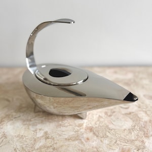 Op de afbeelding: Een gepolijste zilverkleurige metalen olielamp met een druppelvormig lichaam, een gebogen handvat dat omhoog steekt en een zwart accent op de tuit. Het vat heeft een centraal rond deksel met een zwarte knop en rust op kleine, subtiele voetjes.