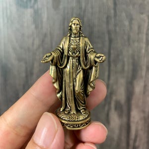 Könnte beinhalten: Eine kleine, metallische, messingfarbene Figur von Jesus Christus, die mit ausgebreiteten Armen steht. Die Figur zeigt ein detailliertes Gewand und ein sichtbares Herz-Jesu-Emblem auf der Brust, platziert auf einem runden, texturierten Sockel.