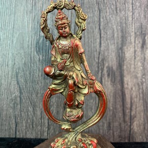 Könnte beinhalten: Detaillierte Statue einer sitzenden Figur mit Heiligenschein, in Rot- und Goldtönen. Die Figur steht auf einem dekorativen Sockel mit aufwendigen Details in Kleidung und Ornamentik. Die Statue ist etwa 15 cm hoch.