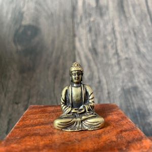 Może przedstawiać: Mała, brązowa statuetka Buddy w pozycji siedzącej medytacji. Figurka stoi na małej, czerwonobrązowej drewnianej powierzchni, z rozmytym drewnianym tłem. Posąg ma szczegółowe rysy twarzy i ubranie.