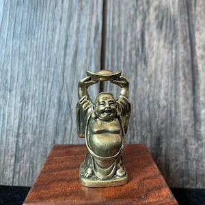 May include: A small, bronze-colored Buddha statue with arms raised, holding a gold ingot. The smiling figure is depicted in traditional robes. The statue is placed on a wooden surface against a gray wooden backdrop. A decorative item.