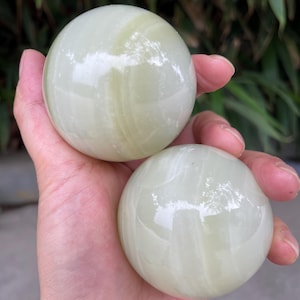 Puede incluir: Dos piedras esféricas pulidas con un acabado liso y brillante. Las piedras son de color verde claro y presentan sutiles patrones de bandas blancas translúcidas en toda su superficie, creando una apariencia veteada en las formas redondas.