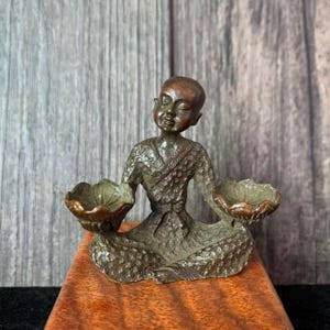 Könnte beinhalten: Bronze-Figur einer sitzenden Person, die zwei lotusförmige Schalen hält. Die Person trägt ein Gewand und sitzt auf einem Holzsockel. Die Figur hat eine detaillierte Textur und ein verwittertes Aussehen.