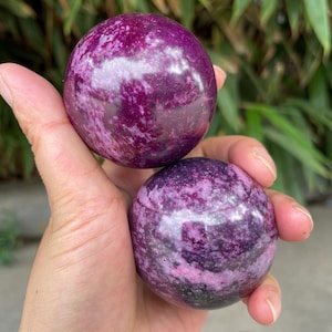 May include: Two polished spherical stones featuring a deep magenta and purple colour palette with mottled white and dark grey mineral inclusions throughout the smooth, rounded surfaces of these two distinct circular mineral specimens.