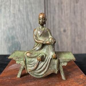May include: A bronze-colored statue of a seated figure in flowing robes, resting on a small bench. The statue has a serene expression and is likely a representation of a spiritual or religious figure. The bench has decorative detailing.