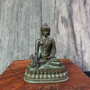 Könnte beinhalten: Eine bronzefarbene Buddha-Statue in sitzender Position. Die Figur hat einen ruhigen Ausdruck und ist mit aufwendigen Details auf dem Gewand und dem Sockel versehen. Die Statue steht auf einer Holzoberfläche.