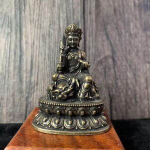 Könnte beinhalten: Eine bronzefarbene Statue einer sitzenden Figur, die eine Krone trägt und einen Stab hält. Die Figur sitzt auf einem Lotusblüten-Sockel, mit komplizierten Details und einem ruhigen Ausdruck. Die Statue ist etwa 10 cm hoch.