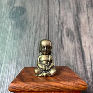 May include: A small, brass-colored statue of a seated monk in a meditative pose. The figure is on a small, wooden stand with a rich, brown finish. The monk's eyes are closed, and the hands are resting in the lap.