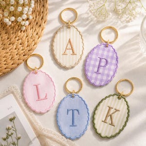 May include: Five oval-shaped fabric keychains with scalloped edges and gold-tone metal rings. Each keychain features a single embroidered letter: A on a cream striped background, P on a purple gingham, L on solid pink, T on solid blue, and K on green striped.