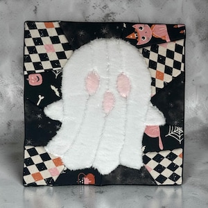 May include: A square fabric wall hanging featuring a central white fuzzy ghost applique with pink eyes and mouth. The background is a patchwork of black and white checkered fabric, black fabric with stars, and patterns of pumpkins, cats, and spiderwebs.