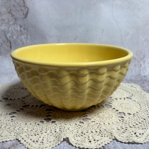 May include: A yellow ceramic bowl with a wavy pattern around the exterior. The bowl sits on a cream-colored, lace doily. The background is a mottled gray and white surface.