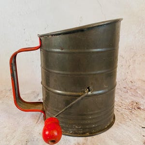 May include: Vintage metal flour sifter with a red handle and a red wooden knob. The sifter is cylindrical with a dark gray finish and visible text. A wire mechanism extends from the side to the red knob. The handle is a bright red.