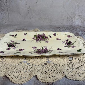 May include: A rectangular, off-white ceramic serving tray with a floral pattern of purple violets. The tray has a scalloped edge with gold trim and is resting on a cream-colored, crocheted doily.