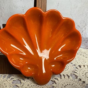 May include: An orange ceramic shell-shaped dish with a glossy finish. The dish has a scalloped edge and a fan-like design, with three small indentations at the base. The dish is likely used for serving or display.