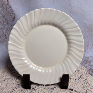 May include: A white ceramic plate with a wavy, ridged rim. The plate is round with a smooth, central surface. The plate is displayed on a dark brown stand, against a textured background.