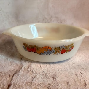 May include: Off-white, round serving bowl with two handles. The bowl features a colorful fruit design around the exterior, including red tomatoes, orange oranges, and purple grapes. A vintage kitchenware item.