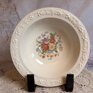 May include: A white ceramic serving bowl with a floral design in the center. The bowl has a decorative border with a grape vine pattern. The floral design includes red, purple, and blue flowers. The bowl is on a dark brown stand.