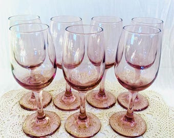 May include: A set of eight vintage pink stemmed wine glasses. The glasses have a delicate, rounded bowl and a slender stem, with a matching pink base. The glasses are arranged on a cream-colored, lace doily.