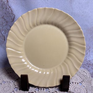 May include: A cream-colored ceramic plate with a ridged, wavy edge. The plate is round with a smooth center and is displayed on a dark brown stand. The plate's design is simple and elegant, suitable for serving food.
