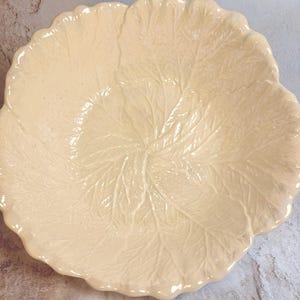 May include: A cream-colored, leaf-shaped ceramic serving bowl. The bowl features detailed veining and a scalloped edge, creating a naturalistic design. The bowl is likely used for serving salads or other dishes.
