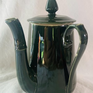 May include: A dark green ceramic teapot with a lid and a curved handle. The teapot has a glossy finish and a spout that curves upward. The lid is topped with a conical finial. The teapot is likely for serving tea or other hot beverages.