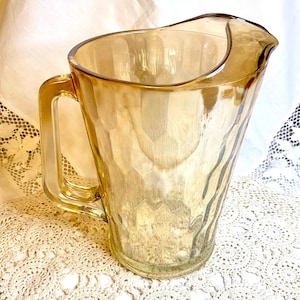 May include: Amber-colored glass pitcher with a textured surface and a rectangular handle. The pitcher has a curved spout and is likely vintage. The pitcher is sitting on a cream-colored, crocheted doily.