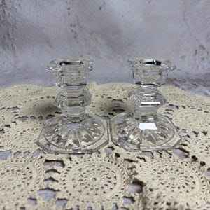 May include: Two clear, faceted glass candle holders. Each has a hexagonal base and a decorative, geometric design. The candle holders are displayed on a cream-colored, crocheted doily, with a neutral background.