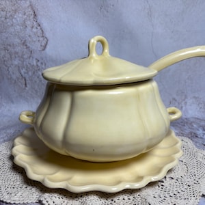 May include: A pale yellow ceramic soup tureen with a lid and a handle, resting on a matching scalloped plate. The tureen has two small handles on the sides. The lid has a loop handle. The set is designed for serving soup.
