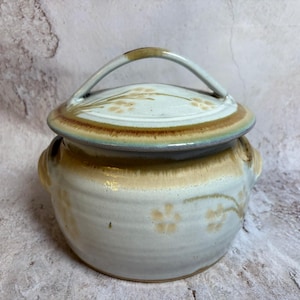 May include: A ceramic lidded jar with a handle. The jar is off-white with brown and tan accents. Floral designs are painted on the lid and the body of the jar. The handle is arched and brown.
