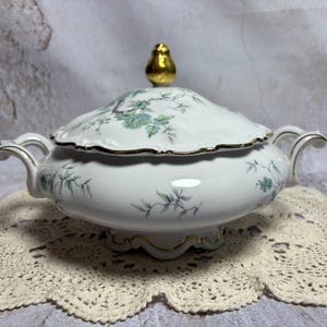 May include: White porcelain serving dish with lid, featuring a floral pattern in shades of green and gold accents. The lid is topped with a gold finial. The dish has two handles and sits on a decorative base. A vintage piece.