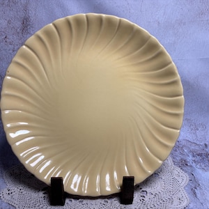 May include: A cream-colored ceramic plate with a spiral design. The plate is round with a raised, ridged pattern radiating from the center. It is displayed on a dark brown stand against a textured background.