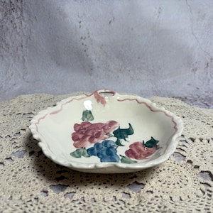 May include: A small, off-white ceramic dish with a scalloped edge and a handle. The dish is decorated with hand-painted flowers in shades of pink, blue, and green. A pink border accents the rim. The dish is resting on a cream-colored lace doily.