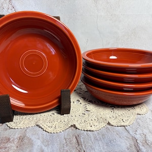 May include: A set of six orange-red ceramic bowls. One bowl is displayed on a wooden stand, while the other five are stacked. The bowls have a circular design with concentric rings and a glossy finish. The bowls are approximately 8 inches in diameter.