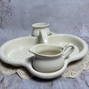 May include: A white ceramic serving set with a woven texture design. The set includes a tray with two compartments, a small pitcher, and a handle. The set is placed on a white lace doily.