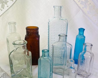 May include: A collection of antique glass bottles in various shapes and sizes. The bottles are clear, brown, and blue, with some featuring embossed details. The arrangement includes tall, rectangular, and small, rounded bottles, showcasing vintage apothecary or medicine bottle designs.
