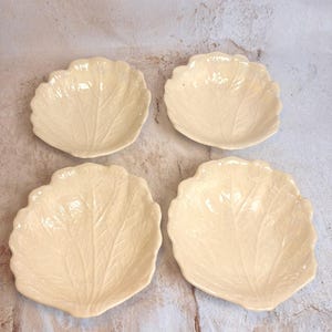 May include: Four off-white, leaf-shaped ceramic dishes. Each dish has a detailed vein pattern and a scalloped edge. The dishes are arranged on a neutral-toned surface, showcasing their naturalistic design.
