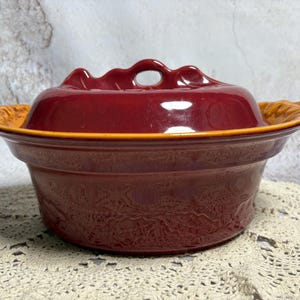 May include: A maroon ceramic casserole dish with a lid. The lid has a handle and sits atop the dish. The dish has an orange rim and a decorative floral pattern. The dish is on a cream-colored lace doily.
