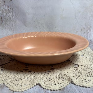 May include: A peach-colored ceramic bowl with a ridged rim. The bowl sits on a cream-colored, crocheted doily. The background is a mottled gray and white surface.