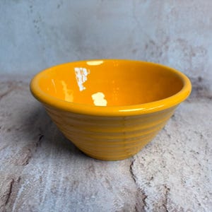 May include: A bright yellow ceramic bowl with a rounded shape and a slightly flared rim. The bowl has a smooth, glossy finish and a series of horizontal ridges around the exterior. The bowl is empty and sits on a textured surface.