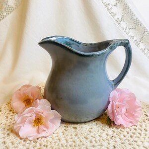 May include: A blue-gray ceramic pitcher with a curved spout and handle. The rim has a darker blue glaze. Three pink roses rest on a cream-colored, crocheted doily, creating a rustic, vintage aesthetic.