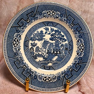 May include: A blue and white ceramic plate with a gold rim. The plate features a detailed landscape scene in the center, surrounded by a patterned border. The plate is displayed on a gold stand, showcasing its intricate design and classic style.