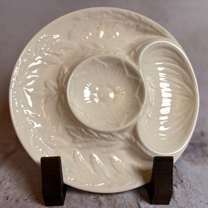 May include: Off-white ceramic serving dish with a floral and leaf design. The dish has a central bowl and a curved section, ideal for dips and snacks. It is displayed on a wooden stand.