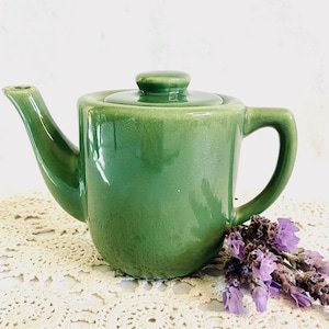 May include: A small, green ceramic teapot with a matching lid. The teapot has a curved spout and a rounded handle. A sprig of purple flowers rests near the base of the teapot. The teapot is sitting on a white, lace doily.