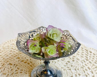 May include: A silver-colored, pedestal-style dish with an ornate, openwork design. The dish holds several green and purple flowers. The dish is sitting on a cream-colored, crocheted surface.