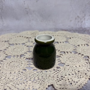 May include: A small, dark green ceramic jar with a white interior and a rounded shape. The jar has a textured surface and a small opening. The jar is sitting on a cream-colored, lace doily.