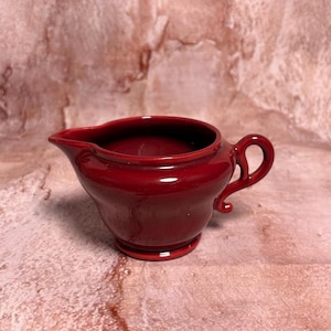 May include: A small, glossy, red ceramic creamer with a spout and a curved handle. The creamer has a rounded body and sits on a small, circular base. The background is a mottled, pinkish-brown color.